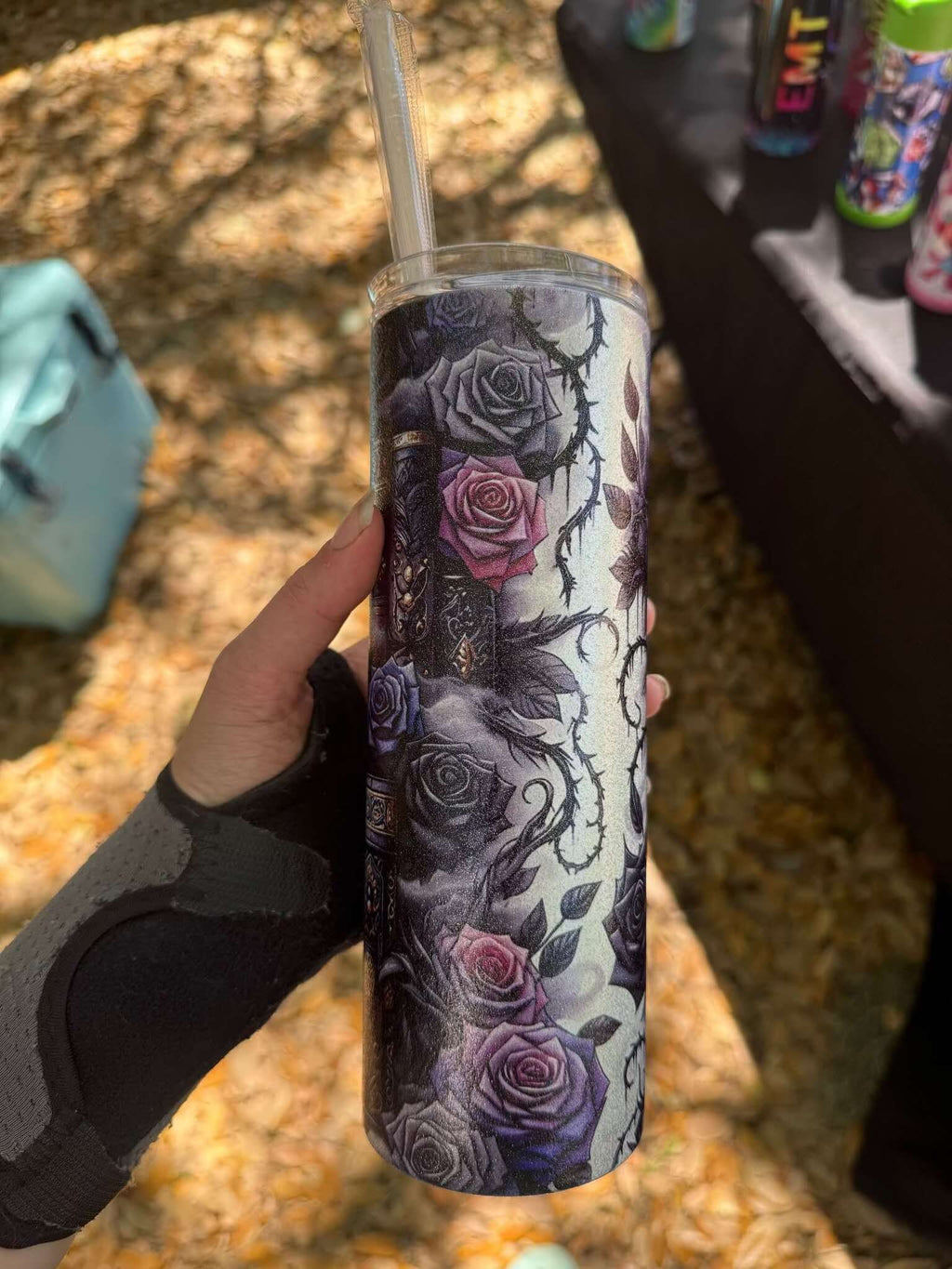 A hand in a wrist brace holds the Dark Book Lover 20oz Skinny Tumbler by Becca's Banging Designs LLC, featuring dark roses, skulls, and barbed wire. A clear straw tops the design, with softly blurred leaves in the background.