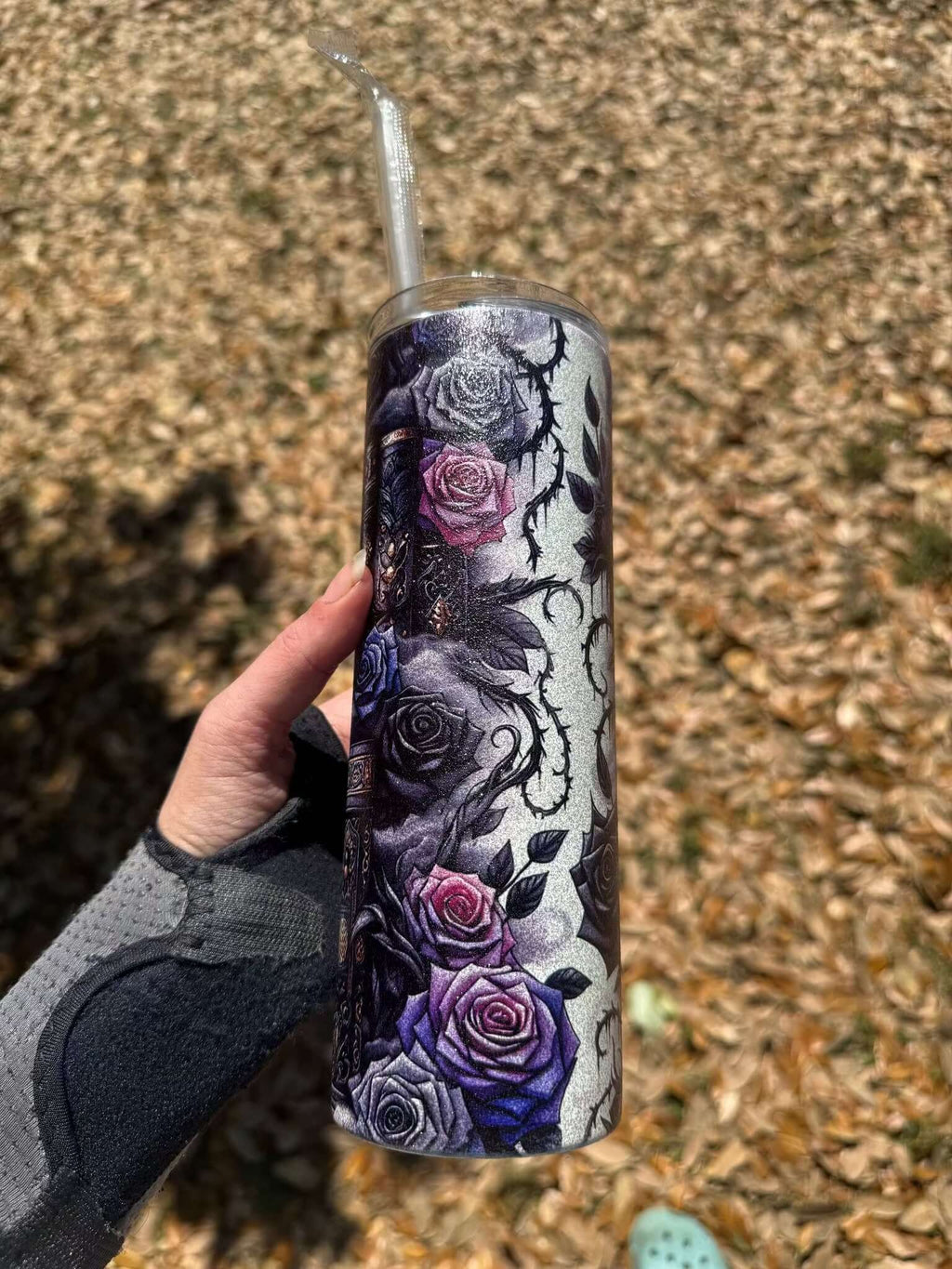 A hand in a gray wrist brace holds the Dark Book Lover 20oz Skinny Tumbler by Becca's Banging Designs LLC, featuring purple and pink roses, thorns, and skulls. Ideal gothic tumbler for book lovers. Background: dry brown leaves.