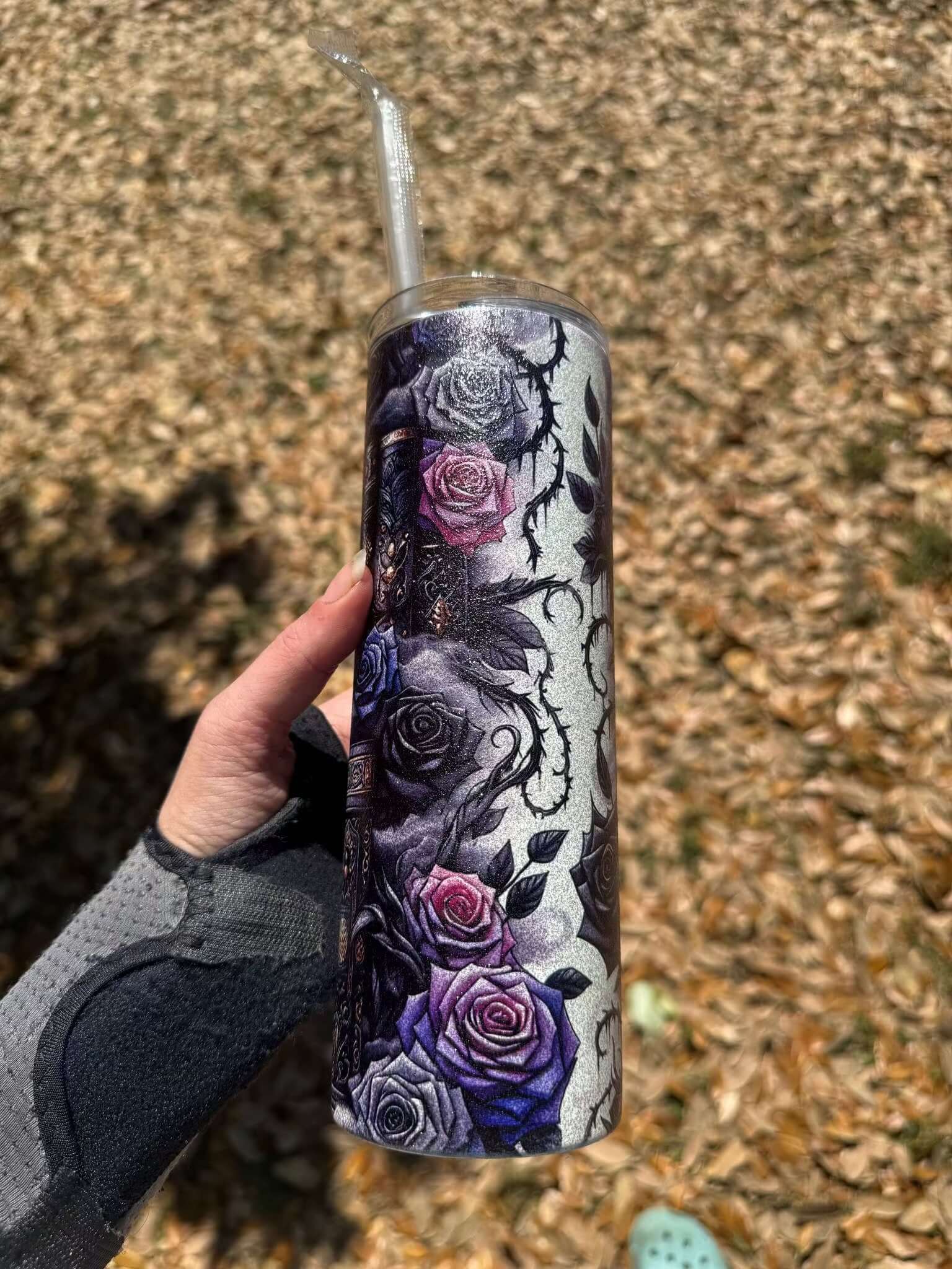 A hand in a gray wrist brace holds the Dark Book Lover 20oz Skinny Tumbler by Becca's Banging Designs LLC, featuring purple and pink roses, thorns, and skulls. Ideal gothic tumbler for book lovers. Background: dry brown leaves.