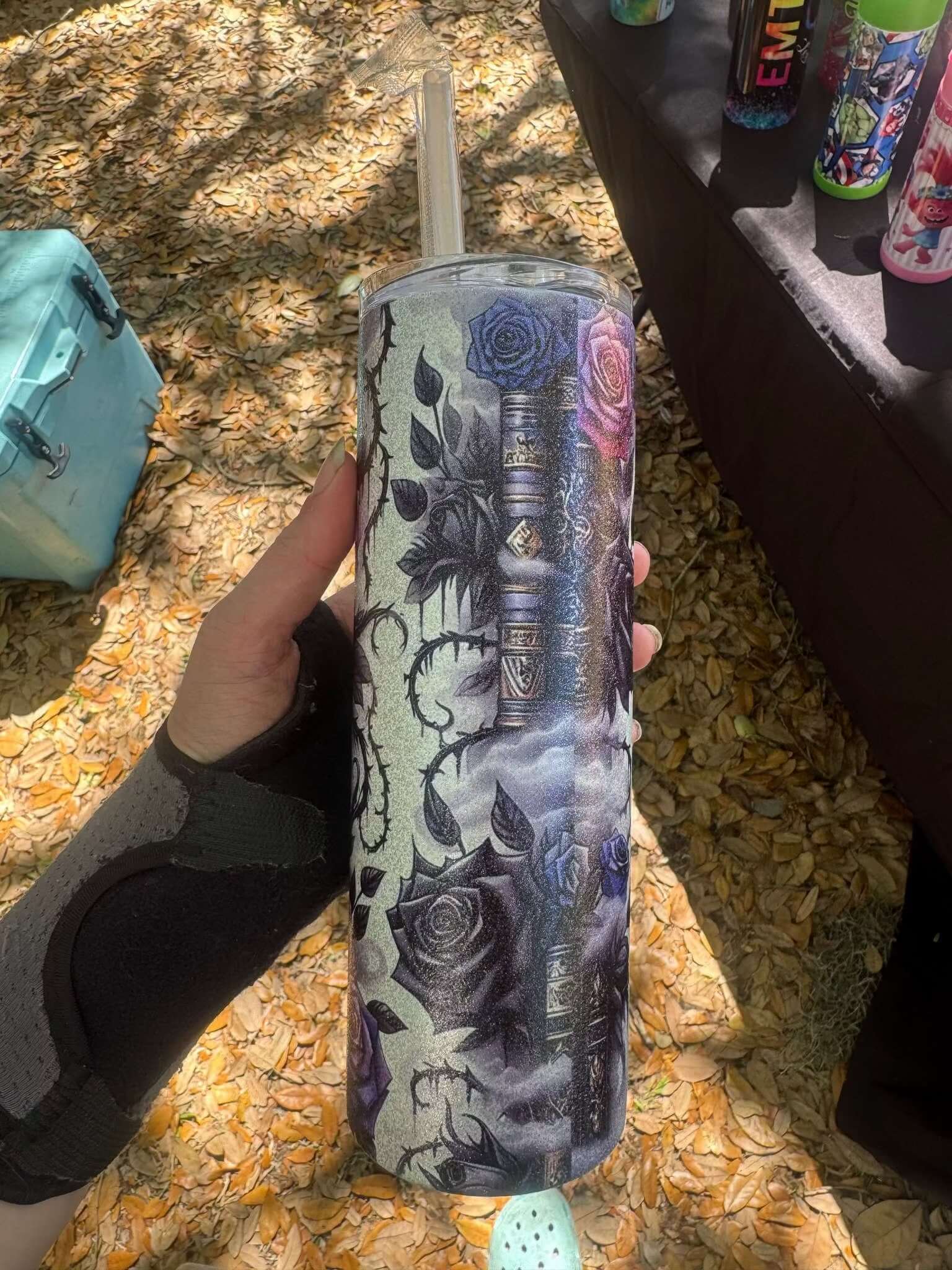 A hand in a black wrist brace holds the Dark Book Lover 20oz Skinny Tumbler by Becca's Banging Designs LLC, featuring dark roses, vintage books, and leaves—a gothic drinkware piece perfect for book lovers. Fallen leaves cover the ground below.