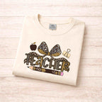Leopard Love Teacher T-Shirt featuring a leopard print bow, apple, pencil, and paperclip design on a soft beige background.